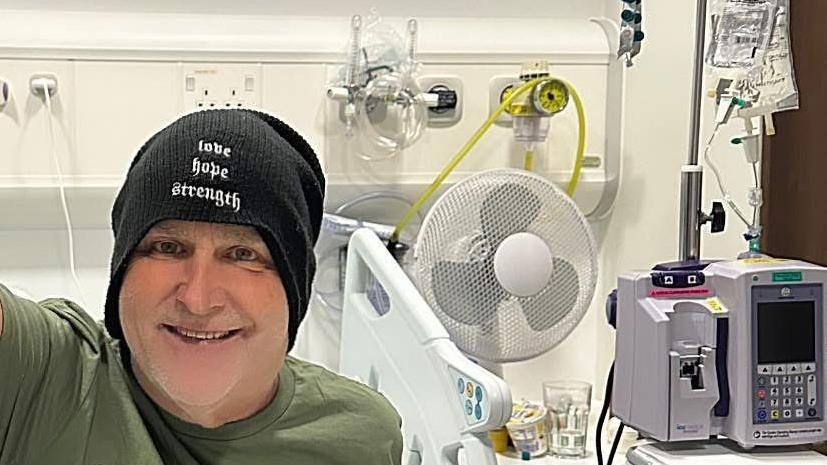 Mike Peters in hospital bed smiling at camera with medical equipment behind him