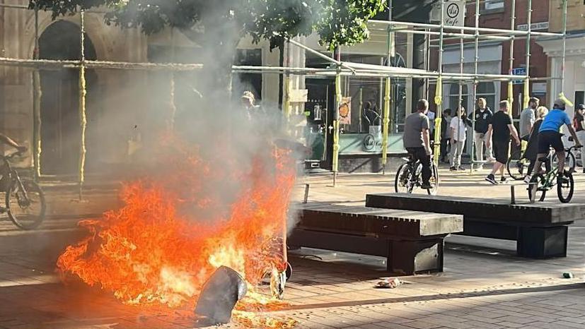 Upturned public bin set alight and raging with fire with people in the background either walking or cycling