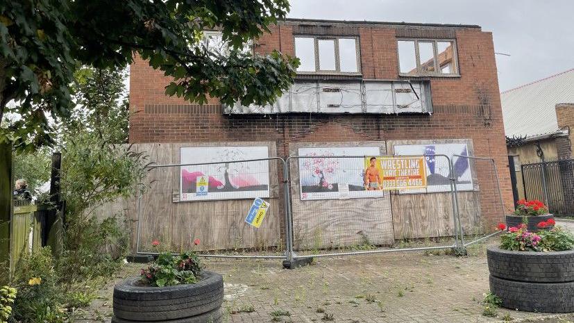 A derelict brick building which boarded up windows on the ground floor and screen off with wire fencing