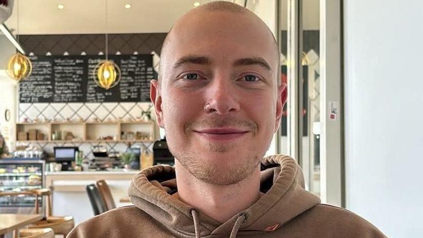 Eli Sweeting. He has close shaven hair and stubble on his chin. He is pictured wearing a brown hoodie and a nose ring. He is looking directly at the camera and smiling.