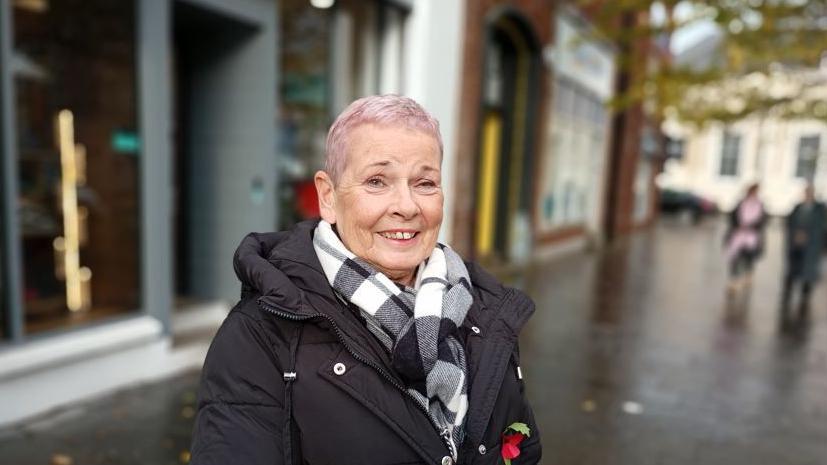Ann McKeown, a woman in black jacket with red poppy and black and white scarf. She has pinkish hair.