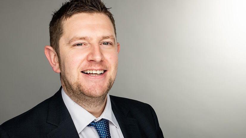 A man with dark hair, swept to his right side and with dark stubble, smiles at the camera. He wears a dark suit, white shirt and blue tie with white dots on it. Behind him is a grey wall.