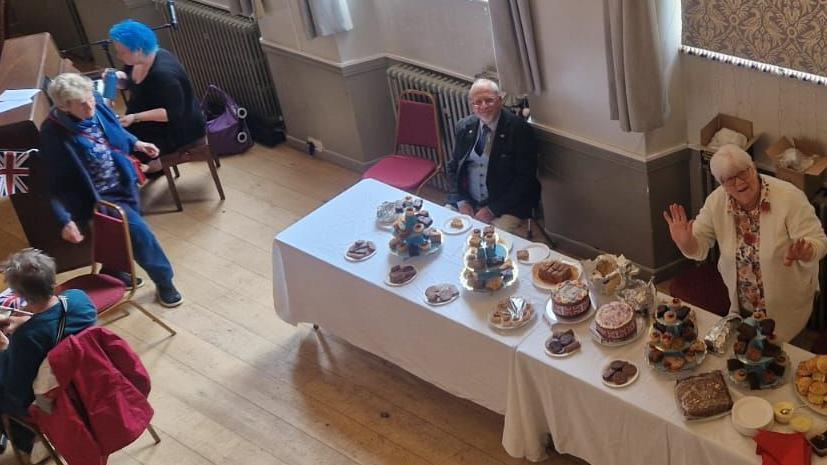A view looking down into a hall ahead of an afternoon tea. Some people are already sitting at tables and on the right is a serving table full of cakes. A man and woman behind the serving table are looking up and waving