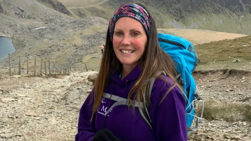 A woman with long brown hair, held back by a colourful headband, stands on a mountain range, wearing a purple top and carrying a blue backpack. She is  smiling at the camera. 