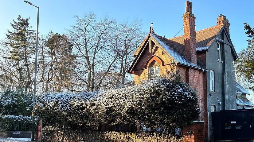 A building with ornamental features and a tall chimney is on the corner of a road and covered with light snow and frost. There are trees with bare branches around and there is a tall hedge outside the property.