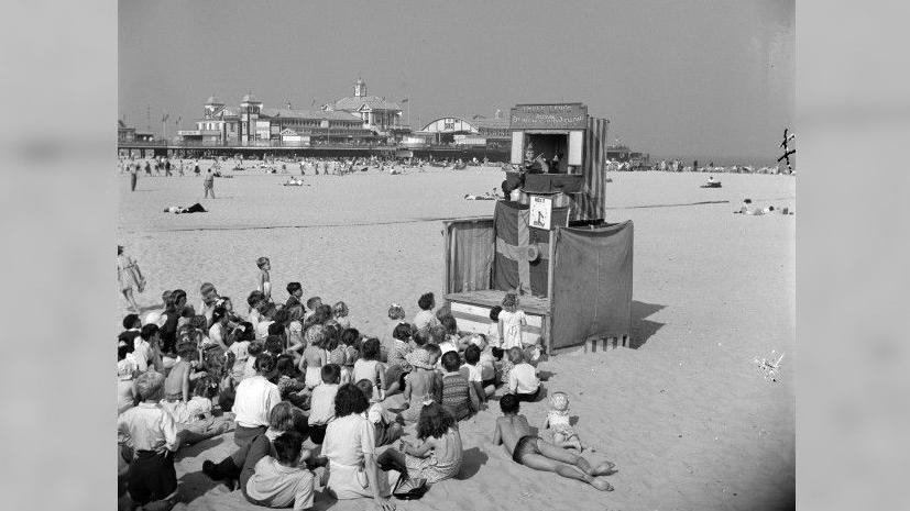A cluster of children sit on the sand to watch Uncle Lew's Punch & Judy Show. Some sprawl on the sand in their swimming costumes. The Britannia Pier can be seen across the beach.