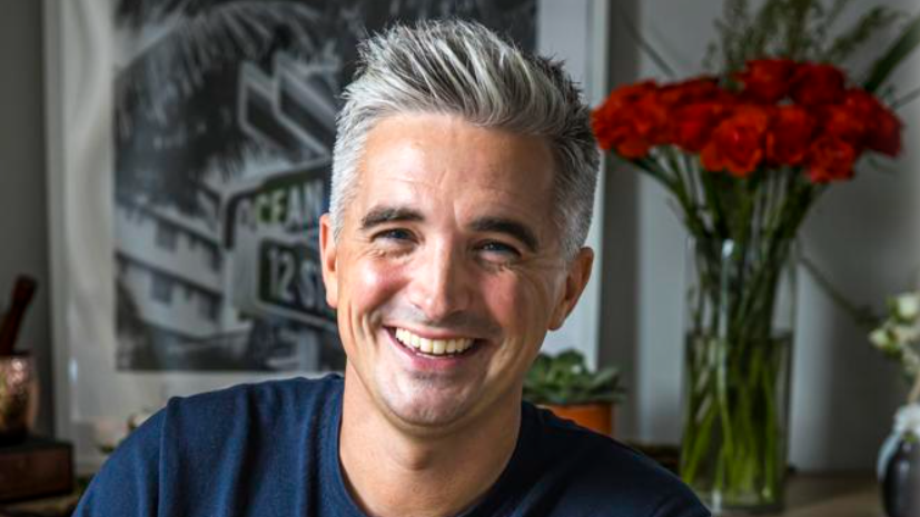 Donal Skehan has salt and pepper hair, parted to the side and raised from his forehead. He is wearing a navy t-shirt and a vase of red flowers is behind where he is sitting