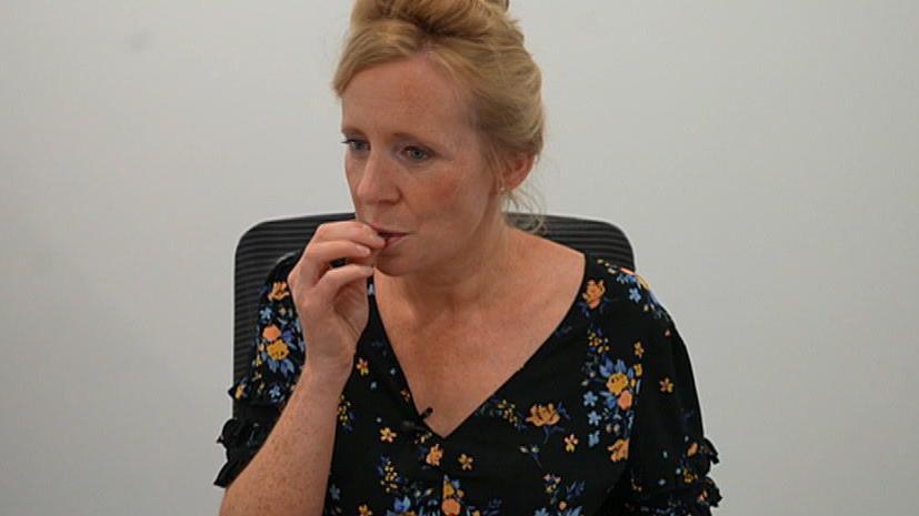 A woman with auburn hair in a top bun hair style tastes a piece of chocolate. She is sitting on a chair and is wearing a black dress, which has blue and orange flowers on it.