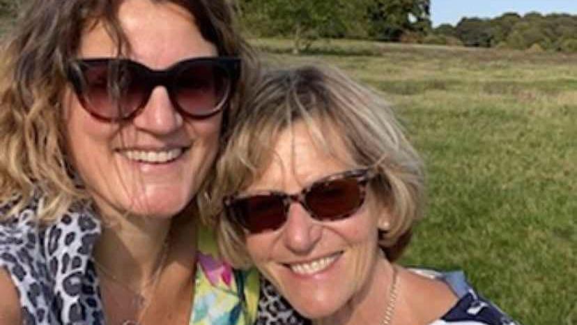 Laura and Liz are hugging, looking at the camera and smiling. They are standing in a field, with trees in the background. Laura has light brown curly hair and wears a grey and black cheetah print scarf loosely around her neck and large sunglasses. Liz has short blonde hair which is parted to the side and a side fringe, she wears tortoiseshell sunglasses, a chain necklace and a blue and white blouse.