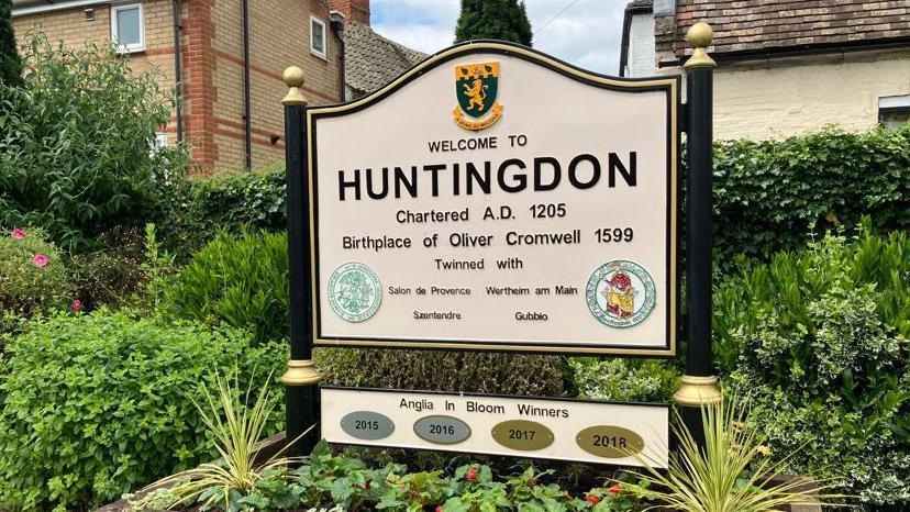 A sign which says 'Welcome to Huntingdon' Birthplace of Oliver Cromwell. 
4 other signs say 'Anglia in Bloom winners' 2015, 2016, 2017, 2018.