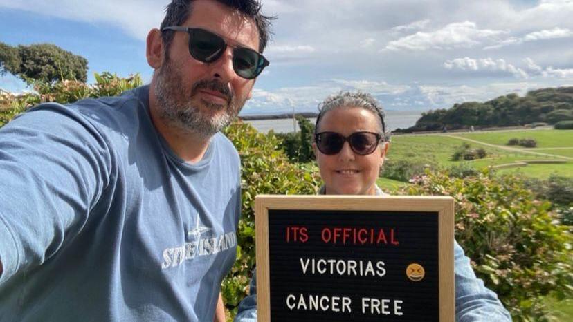 Victoria is holding a sign which reads "It's official, Victoria's cancer free". She and her husband are wearing sunglasses. In the background are The Knap gardens and Old Harbour in Barry.