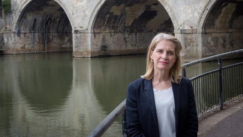 Liberal Democrat MP for Bath Wera Hobhouse. She is wearing a black blazer, a white t-shirt and gold hoop earrings. She is standing beside the water in Bath in front of three tunnels.