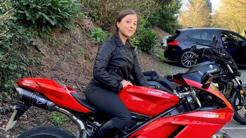 Rakela Kalemi sitting on a red Ducati motorbike in a car park set within woodland