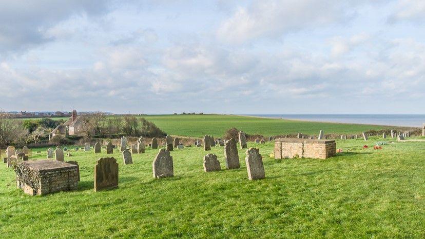 A view of a grass graveyard with a number of grave stones. In the distance is the sea, and a house. 