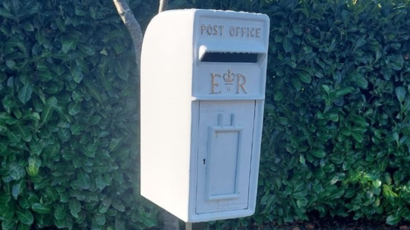 A postbox painted white with gold lettering. Behind it is a hedge.