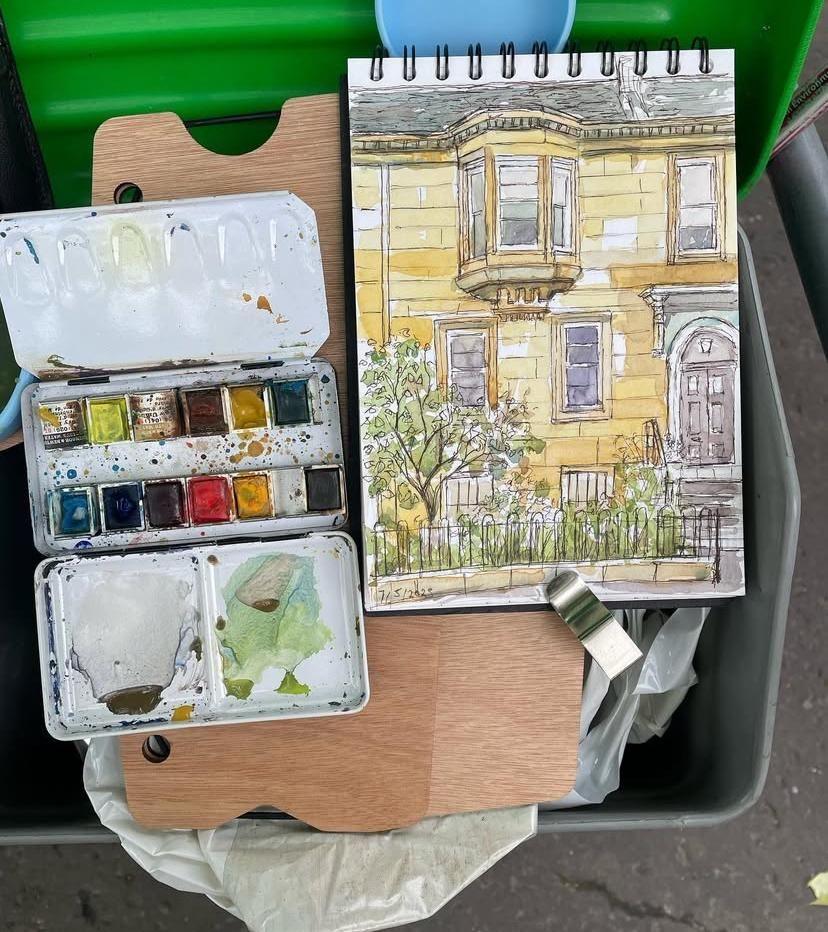 A photograph of a watercolour paint palette and sketchbook with a painting of a tenement flat in Glasgow's west end resting on the top of a street sweeping cart.