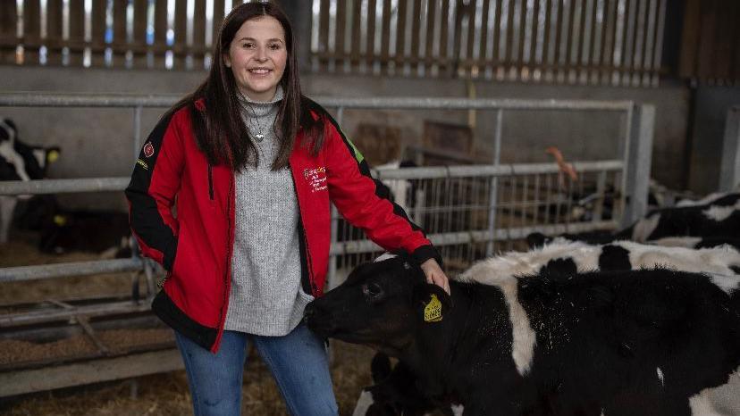 Sara Roberts, 22 - agriculture lecturer at Coleg Sir Gâr - standing with a calf in a cattle shed, wearing a red coat over a grey jumper and jeans.  