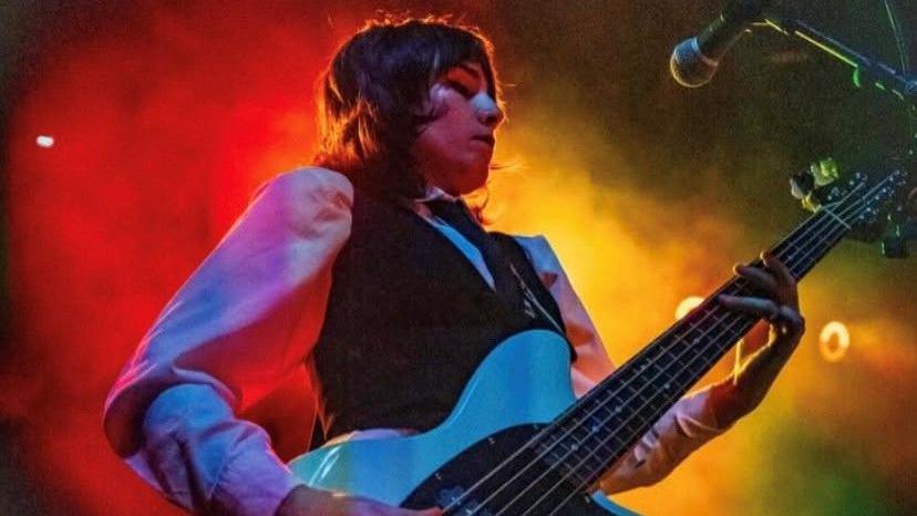 Emer McLaughlin has short brown hair and wears a waistcoat, tie and white shirt.  She holds a guitar in her hands while the background is full of coloured smoke. 