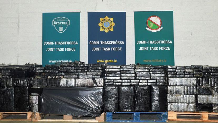 Picture issued by the police showing the recovered cocaine shipment. Piled on wooden pallets, small rectangular blocks are wrapped in black plastic. They are in front of a white wall with posters showing the logos of the Irish authorities.