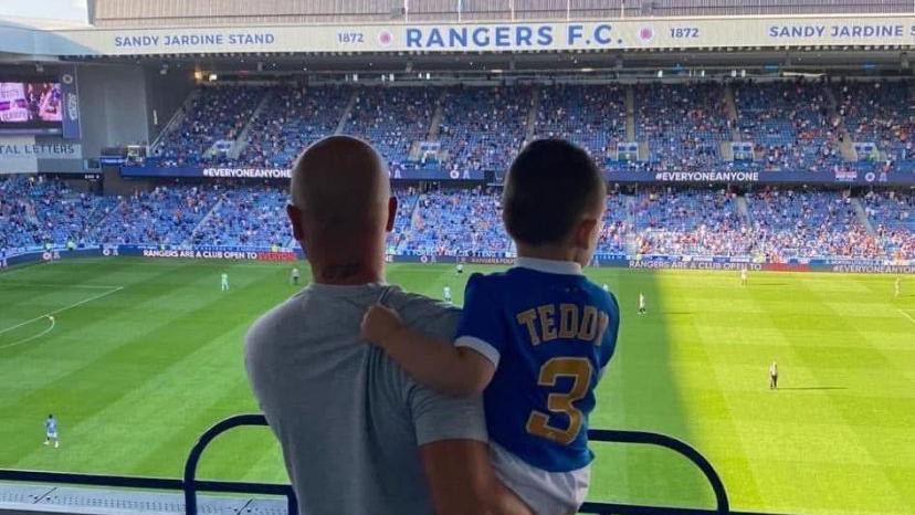 A man holding his son, photographed from behind, looking at the pitch at Ibrox stadium in Glasgow. The man has a shaved head and is wearing a white t-shirt, The boy is wearing a blue Rangers home top with "Teddy 3" on the back of it.