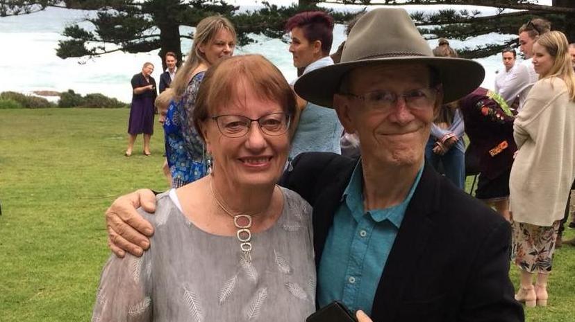 Geraldine has short hair and is wearing glasses. She is wearing a top with feathers on it. Donald is wearing a broad brimmed hat. He has glasses and a dark coloured suit jacket and blue shirt. They are at an event and other people stand near the couple.