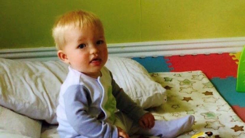 Baby Jackson wearing a baby grow. He has blond hair and looking at the camera