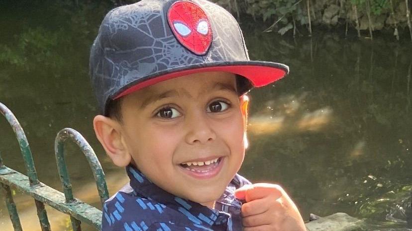 A smiling boy with brown eyes and wearing a Spider-Man cap. He is standing against railings next to a pond.