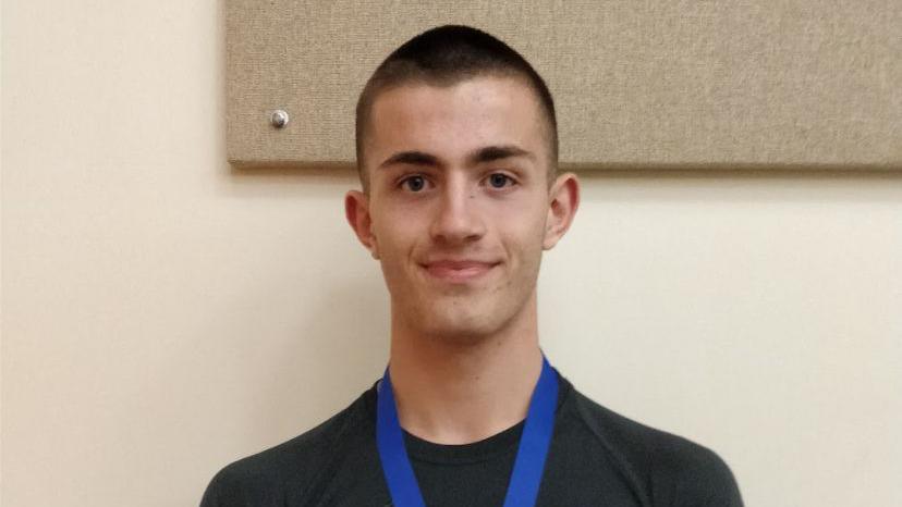 Will Radford is pictured smiling with a medal around his neck. He has short dark hair and wears a black t-shirt with a blue medal ribbon.