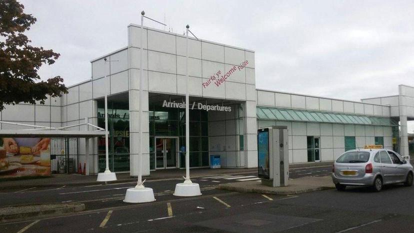 The entrance to City of Derry airport, with a sign above the doors that reads, arrivals and departures. A taxi is parked outside the grey building to the right of the doors