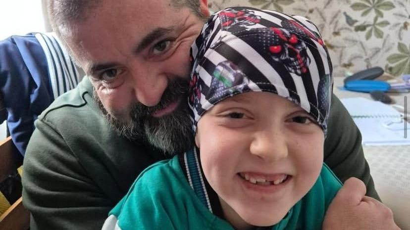 A man with a grey and black beard sits smiling with his arms around a little girl, who is also smiling and wearing a black and white stripy bandana.