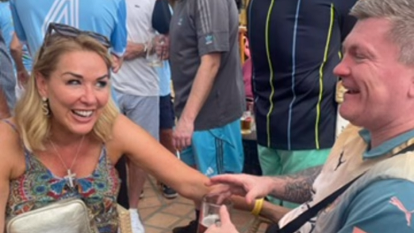 Claire Sweeney sits in a chair opposite Ricky Hatton who is wearing a Manchester City shirt. They are laughing with each other and surrounded by people stood around. 