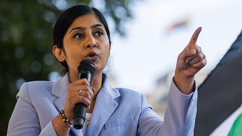 Zarah Sultana, wearing a pale blue suit giving a speech with a microphone.