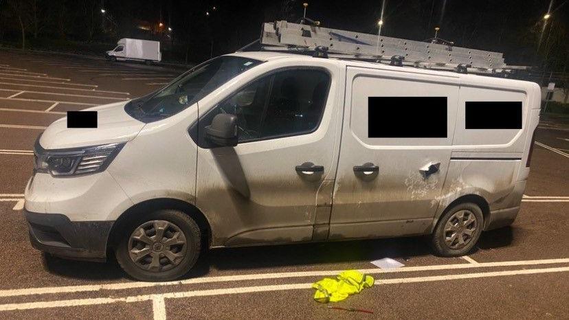 Oliver Powell's white van parked in a car park. It has a ladder attached to its roof. There is a hole in the side door that the thieves drilled. It is night-time.