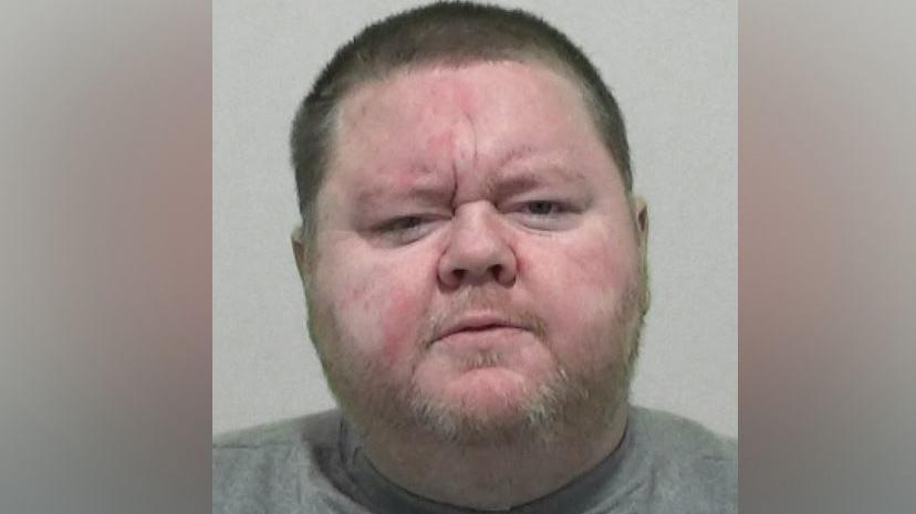 Mugshot of Mark Hogarth. He has a large round head with a stubbly fair beard and short dark hair. he is wearing a grey sweatshirt.