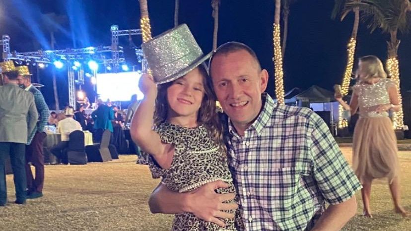 A young Gracie smiles at the camera with her father Simon. Simon has his arm around Gracie and she is wearing a silver top hat and leopard-print dress. Other partygoers appear behind them, some wearing similar silver top hats. 