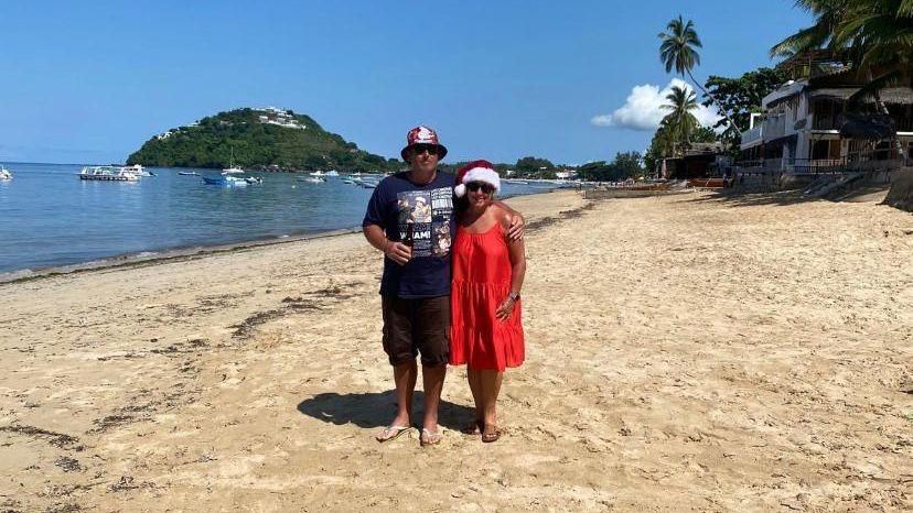 The couple are on a beach in Madagascar, wearing summer clothes and Victoria is wearing a Christmas hat. There are small boats and palm trees in the background.