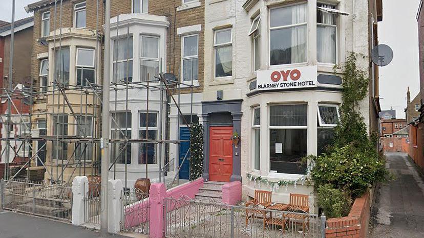 A white guesthouse on the corner of a street with a sign at the front which reads OYO Blarney Stone Hotel. Other properties to the left of the street have scaffolding up.