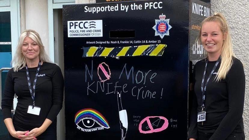 Two women wearing black t-shirts with lanyards around their necks. They are standing either side of a black knife bin, which has drawings and the words "no more knife crime" on the front.