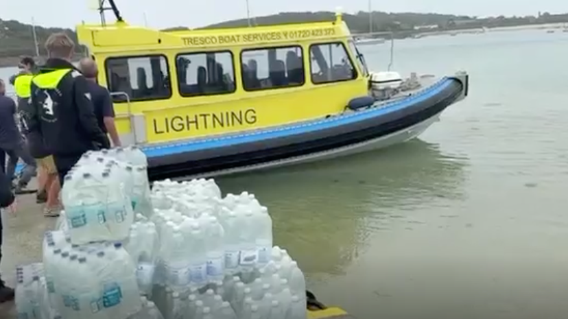 A yellow and blue boat with Lightning written on its side is seen with stacked bottles of water visible in the foreground. Some people are milling around in the background.