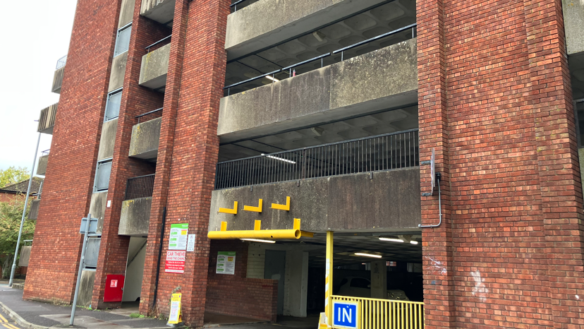 St Stephen's Car Park in Trowbridge. The multi storey car park features discoloured bricks and railings which look old. There is also a blue sign which says 'in' on the road level.