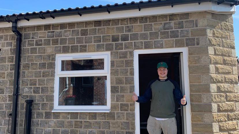 A man stood in the doorway of what appears to be a very small brick house. It is actually an industrial kitchen, which he has built.