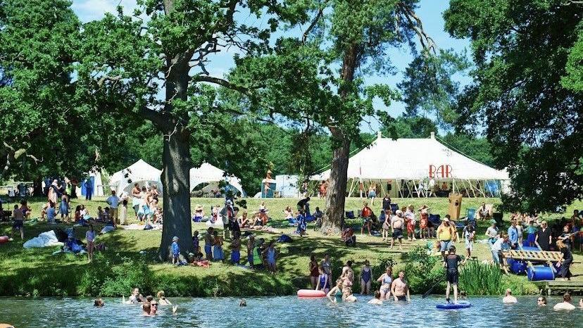 Dozens of people are on a festival site in a field. There are trees next to a body of water and white festival marquees in the background. People are walking, sitting or swimming in the water. One person stands up on a paddleboard