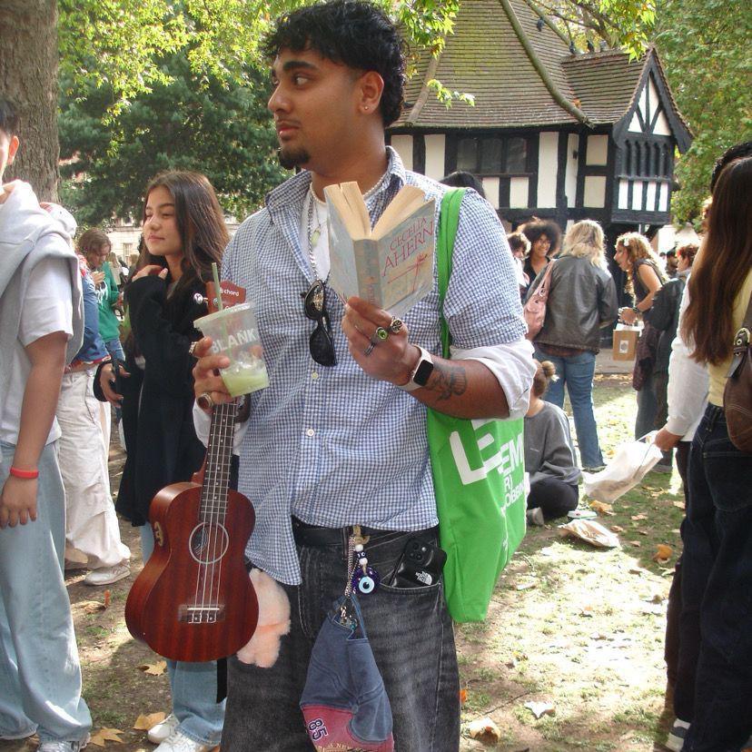 A young man reads PS, I Love You as he stands in a square. He is clutching a ukulele in the other hand and has a tote bag slung over his shoulder. 
