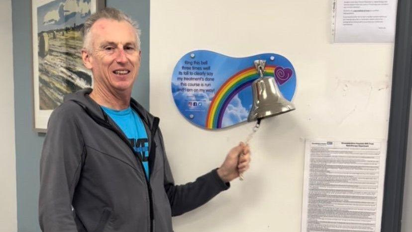 Mr Emerson wearing a blue t-shirt underneath a dark grey zip-up hoodie. He is ringing a silver bell which is attached to the wall in a hospital. He is smiling at the camera but looks thin and tired.