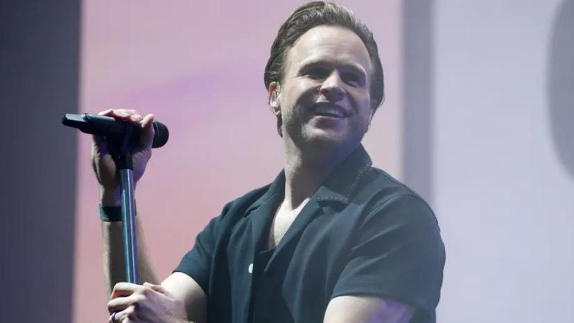 Olly Murs wearing a black button-down top and holding a mic