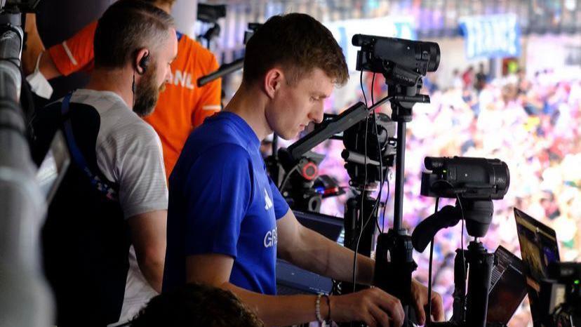 Rhys Llywelyn gyda laptiop a chamera mewn felodrom