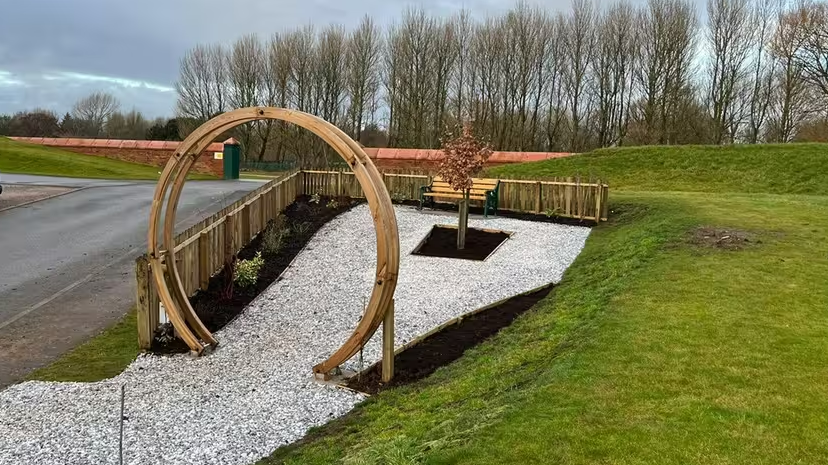 Carlisle United's memorial garden which has a hoop as an entrance with a tree at its centre surrounded by white gravel and a bed of plants to one side with a low wicker fence around it 