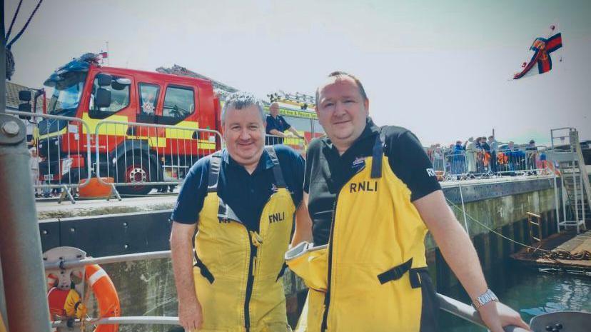 Two men smiling at camera wearing yellow overalls and navy polo shirts. They are standing on the edge of a boat. There is a fire engine in the background on a harbour pier.