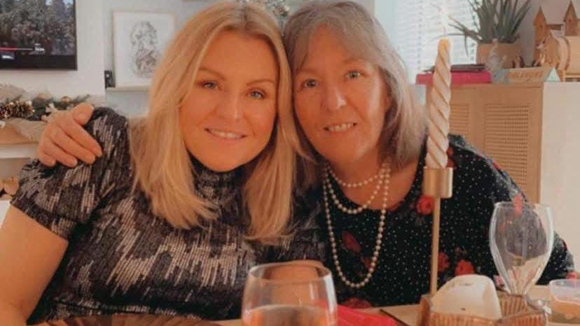 Two women sit at a table with their arms around each other. Both are smiling and looking into the camera. Empty glasses and candles can be seen in front of them.