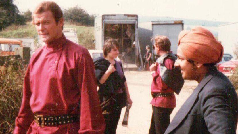 Roger Moore is on the Octupussy film set wearing a red tunic and black belt. He is looking towards the camera while walking away. Looking at him is Del who wears an orange turban. In the background are lorries with equipment and production teams.